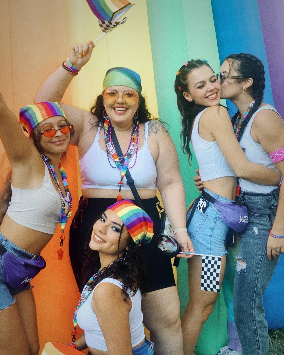 A vibrant group of five friends celebrates against a colorful rainbow backdrop, wearing matching accessories and smiling joyfully.