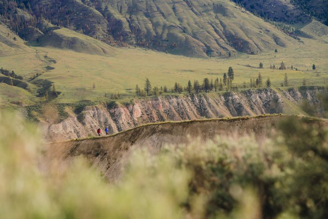 Two hikers traverse a rugged trail along a steep, grassy ridge, surrounded by rolling hills and distant mountains under a clear sky.