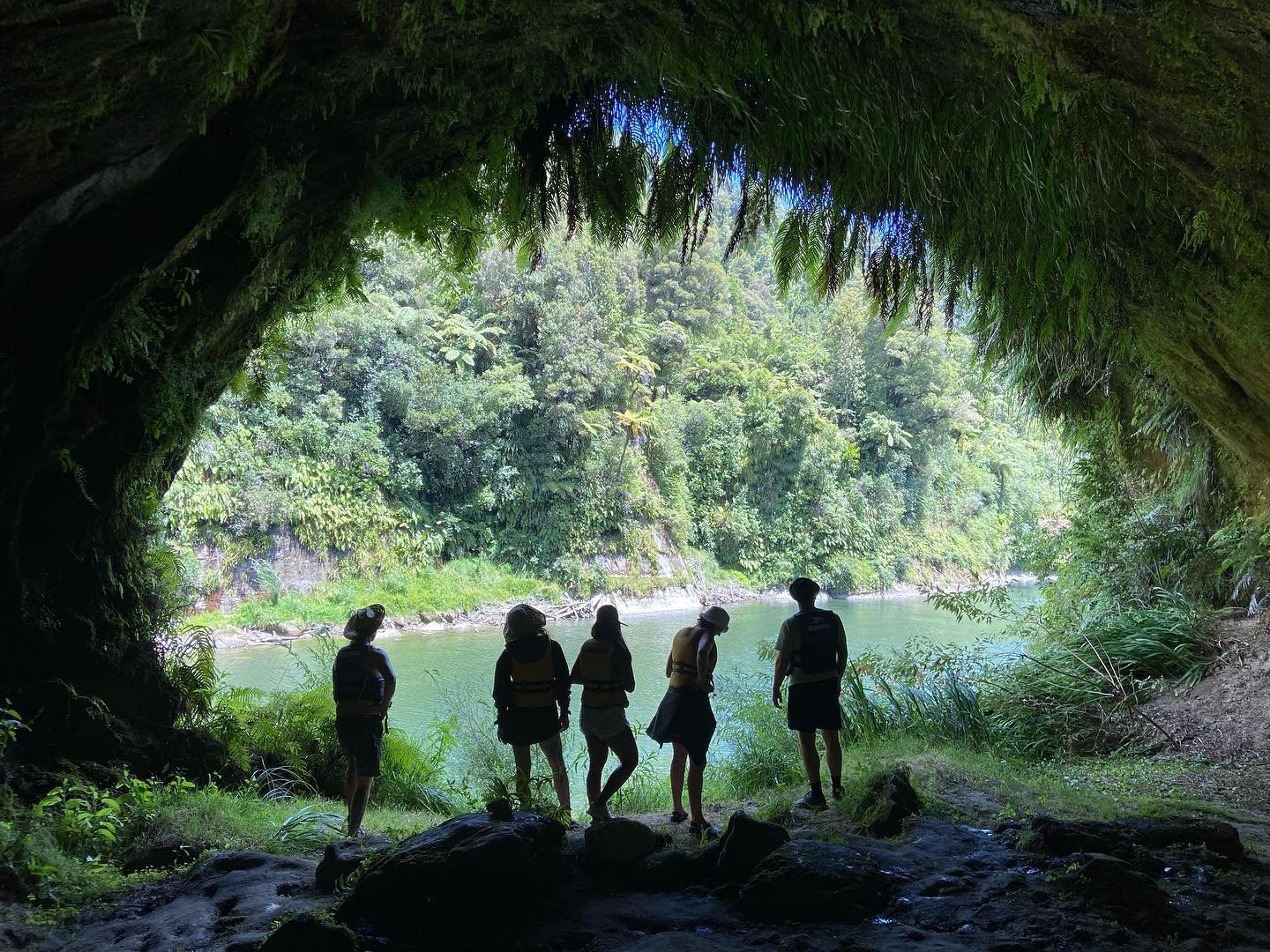 Silhouettes of five people stand at the entrance of a lush cave, overlooking a serene river surrounded by dense greenery.