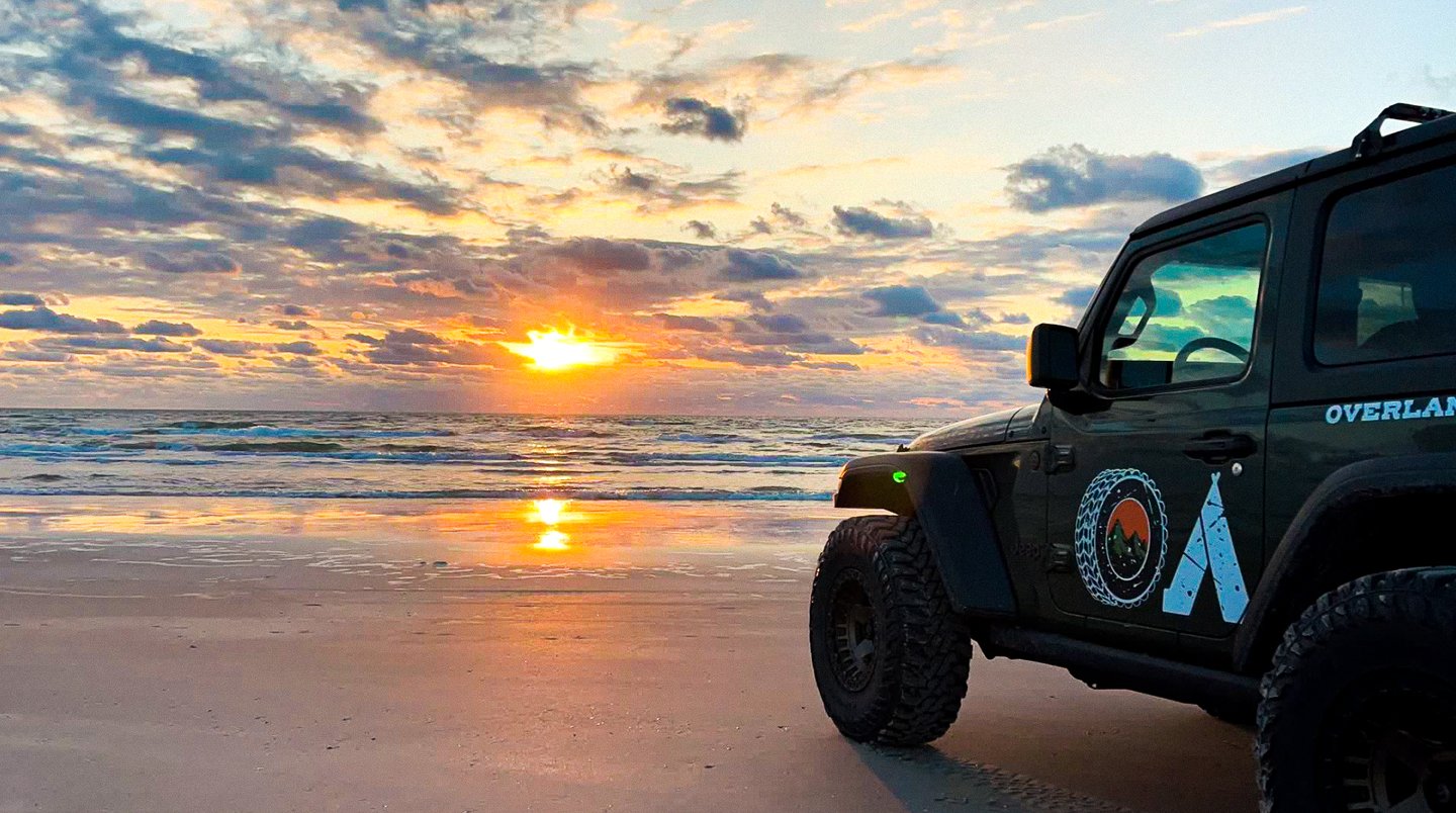 A rugged off-road vehicle parked on a sandy beach at sunset, with vibrant clouds reflecting on the water's surface.