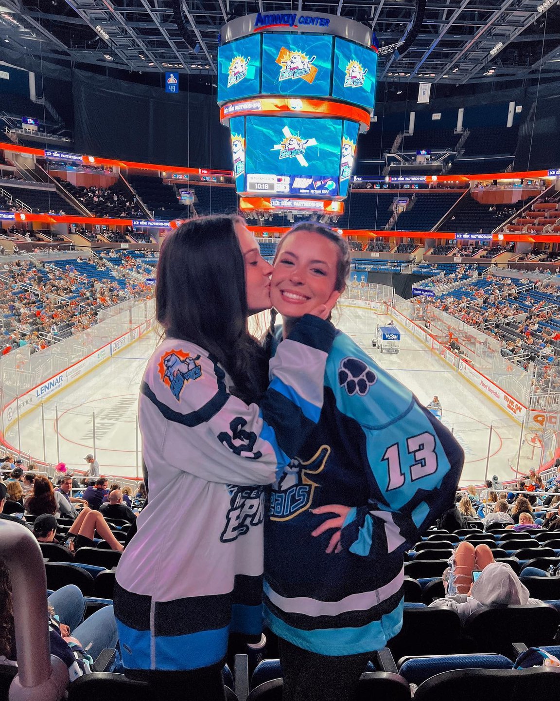 Two women in hockey jerseys smile at the camera in a packed arena, with an ice rink and large scoreboard in the background.
