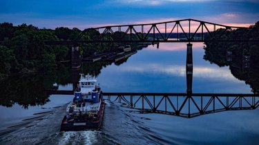 A barge navigates a calm river under a silhouetted bridge at sunset, reflecting vibrant colors on the water's surface. Lush greenery lines the banks.