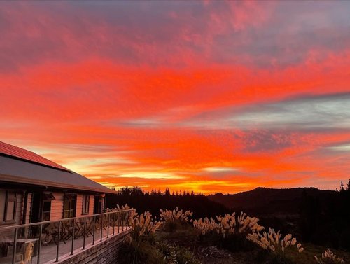 A rustic cabin overlooks a vibrant sunset with hues of orange and pink, framed by lush greenery and distant hills. The scene evokes tranquility.