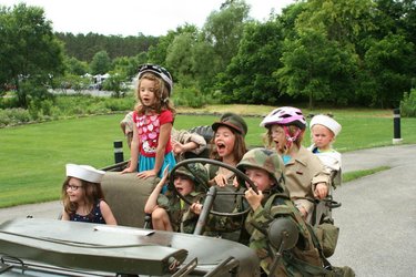 A group of children in military-themed outfits and helmets joyfully pose in an old military vehicle, surrounded by greenery.