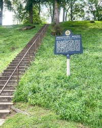 Stone steps lead up a grassy hill to a sign reading "Prehistoric Mound," surrounded by lush greenery and trees.