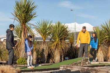 A family enjoys mini-golf surrounded by lush greenery and palm-like plants, with a snow-capped mountain in the background.