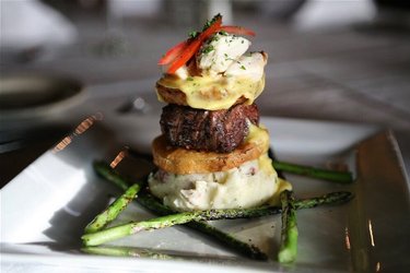 A gourmet dish featuring a stacked presentation of beef, topped with seafood and sauce, surrounded by asparagus on a white plate.