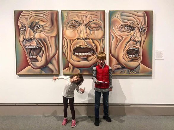 Two children stand in front of three expressive, abstract portraits in a gallery. The artworks depict distorted faces with intense emotions.