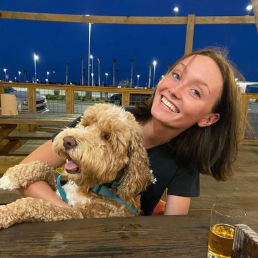 A young woman smiles while holding a fluffy dog at a wooden table outdoors, with a nighttime cityscape in the background.