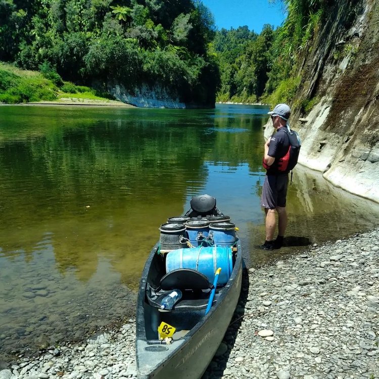 A person stands by a calm river, next to a kayak on a pebbled shore, surrounded by lush greenery and steep cliffs.