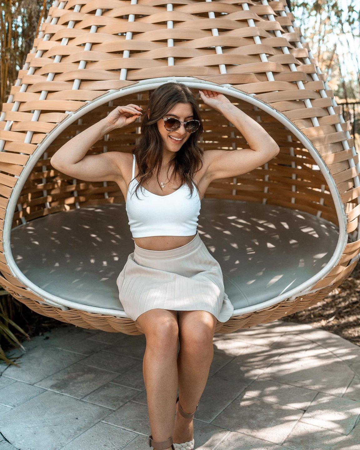 A woman sits in a large, woven hanging chair surrounded by greenery, wearing sunglasses and a casual outfit, enjoying a relaxed moment.