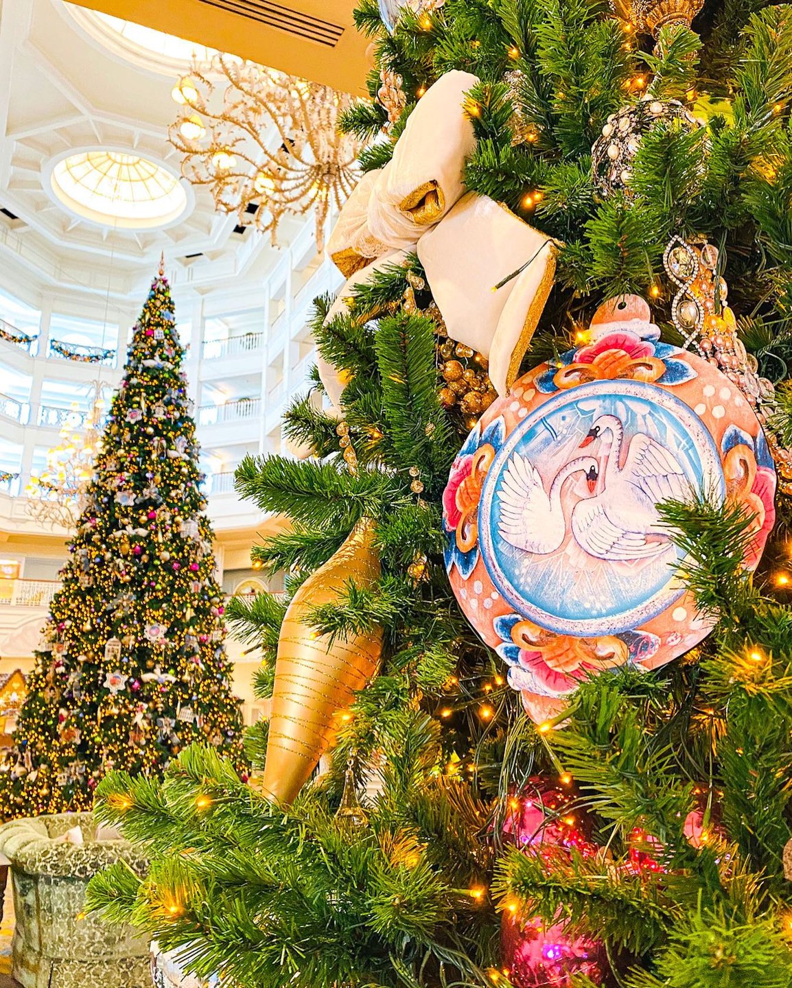A beautifully decorated Christmas tree adorned with colorful ornaments and lights, set in an elegant lobby with a second tree in the background.