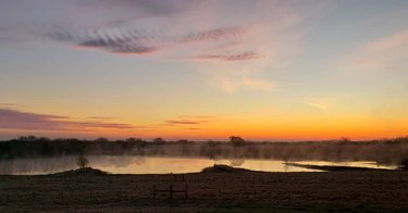 A serene lakeside view at sunrise, with mist rising from the water and colorful clouds reflecting in the sky. Tranquil landscape setting.