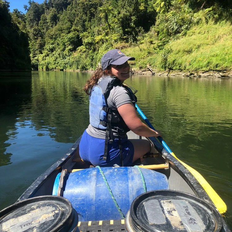 A person paddles a canoe on a calm river surrounded by lush greenery and steep banks, reflecting a serene outdoor adventure.