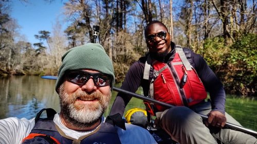 Two men in kayaks on a calm river, surrounded by trees. One man wears a green beanie and sunglasses, while the other sports a red life jacket.