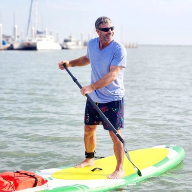 A man stands on a bright yellow paddleboard in calm waters, paddling with a scenic marina in the background. Sunlight reflects off the water.