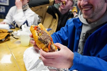 A close-up of a person holding a loaded hot dog with toppings, seated at a table with others enjoying food in a casual setting.