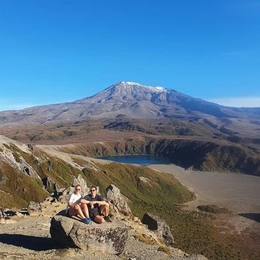 Two hikers sit on a rocky outcrop overlooking a vast valley with a lake, framed by a snow-capped mountain under a clear blue sky.