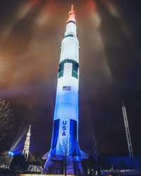 A towering rocket illuminated in red, white, and blue against a night sky, surrounded by trees and structures, showcasing a historic space landmark.