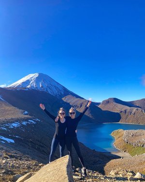 Two hikers stand on a rocky outcrop, arms raised, with a snow-capped mountain and a serene lake in the background. Clear blue skies enhance the stunning landscape.