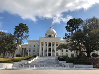 Historic building with a prominent dome and clock, flanked by trees and landscaped steps, under a partly cloudy sky.
