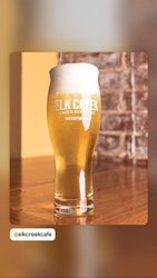 A tall glass of beer with a frothy head sits on a wooden table, featuring the logo of Elk Creek Café & Aleworks. The background shows a rustic brick wall.
