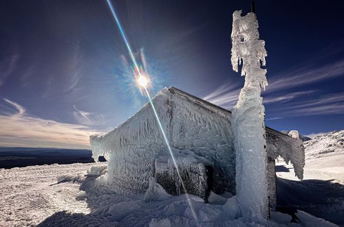 A snow-covered cabin glistens with ice formations under a bright sun, surrounded by a vast, snowy landscape and clear blue skies.