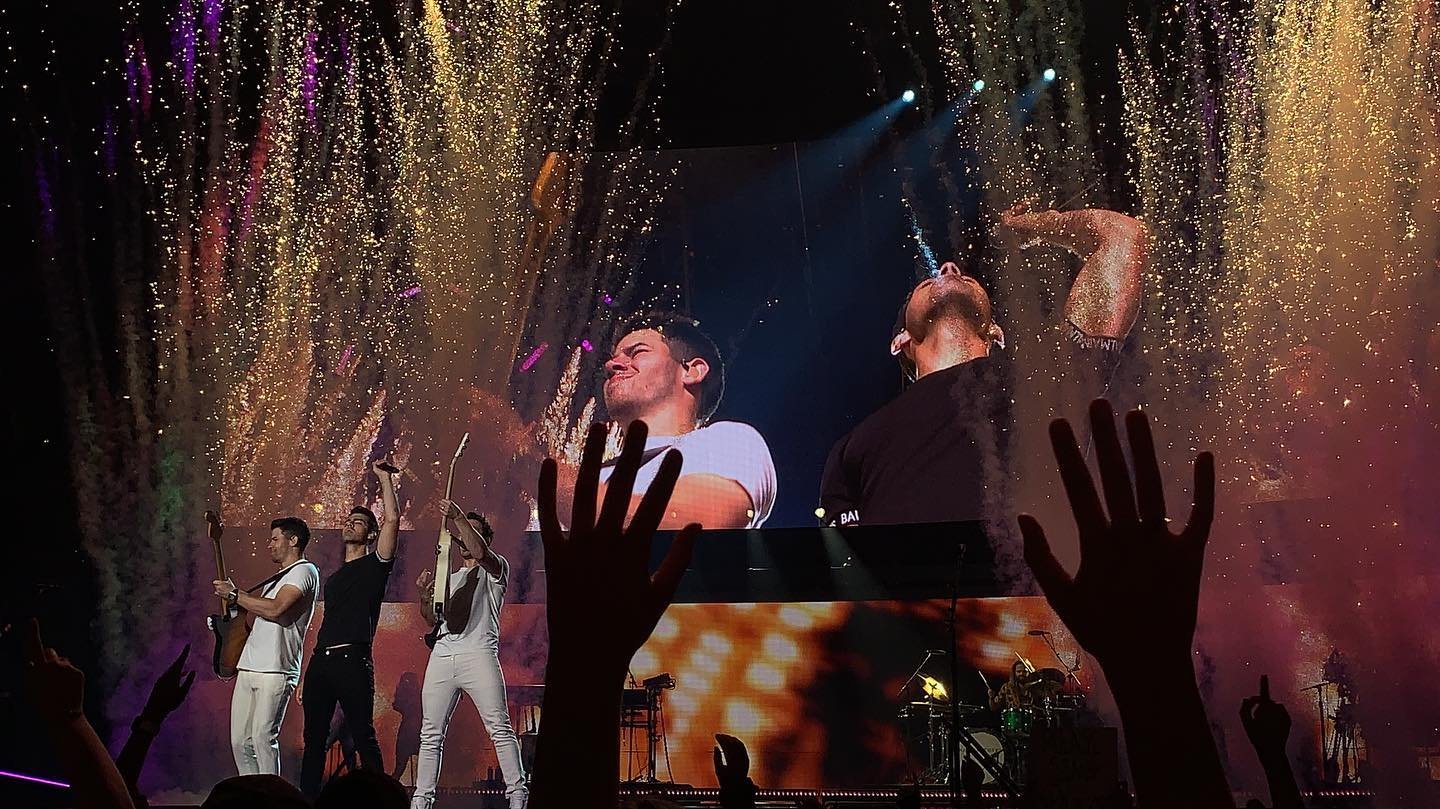 Three performers on stage with raised hands, surrounded by colorful confetti and lights, creating an energetic concert atmosphere.