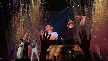Three performers on stage with raised hands, surrounded by colorful confetti and lights, creating an energetic concert atmosphere.