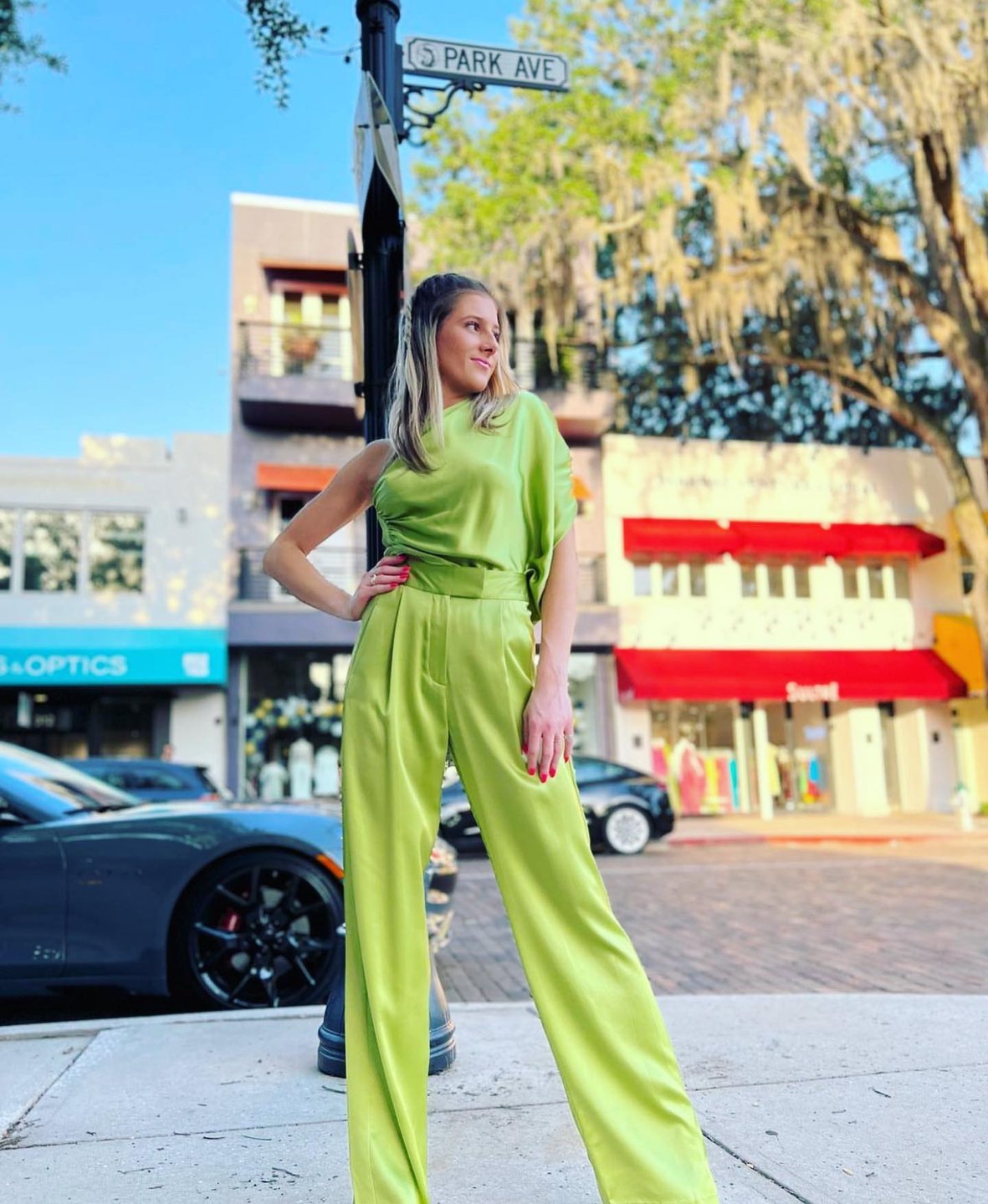 A woman in a vibrant green outfit stands confidently on a city street, with shops and a street sign in the background. Sunlight filters through trees.