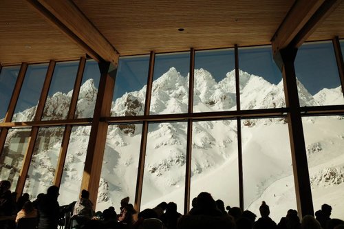 A cozy interior with large windows showcasing snow-covered mountains. People are seated, enjoying the view and atmosphere.
