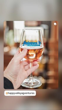 A hand holds a clear wine glass filled with rosé, featuring a logo. The background shows a warm, inviting bar setting.