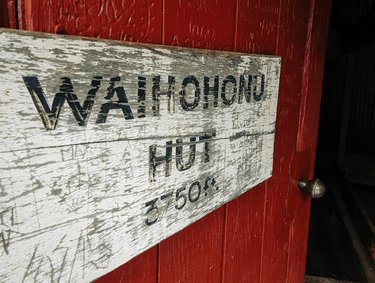 Weathered wooden sign reading "Waihohonu Hut" mounted on a red wall, indicating a rustic accommodation in a natural setting.
