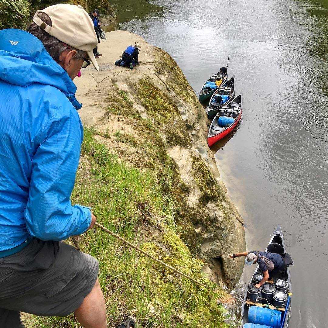 A person in a blue jacket descends a rocky bank to assist others in canoes on a calm river, surrounded by lush greenery.