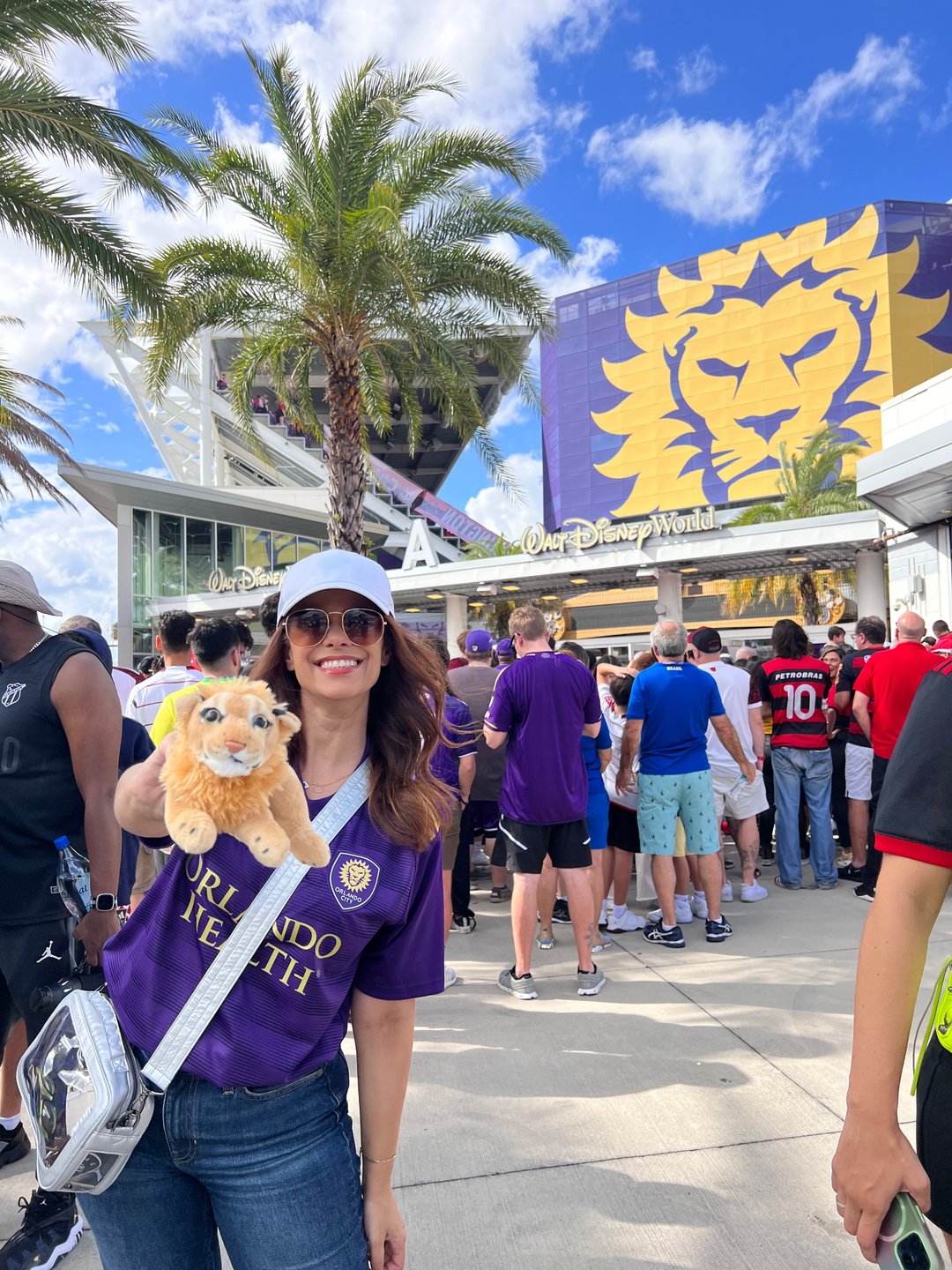 A woman in a purple jersey holds a plush lion toy outside a stadium, surrounded by a crowd and palm trees under a blue sky.