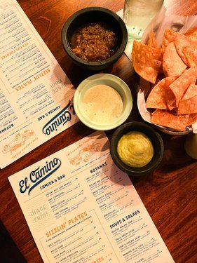 A wooden table displays menus from "El Camino" alongside bowls of salsa, guacamole, and creamy dip, with tortilla chips in a basket.