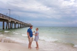 A couple stands on a sandy beach, near gentle waves, with a wooden pier in the background under a cloudy sky.