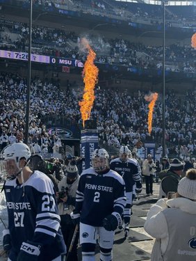 Ice hockey players in blue and white uniforms enter a stadium filled with cheering fans. Flames erupt from pillars, enhancing the excitement.