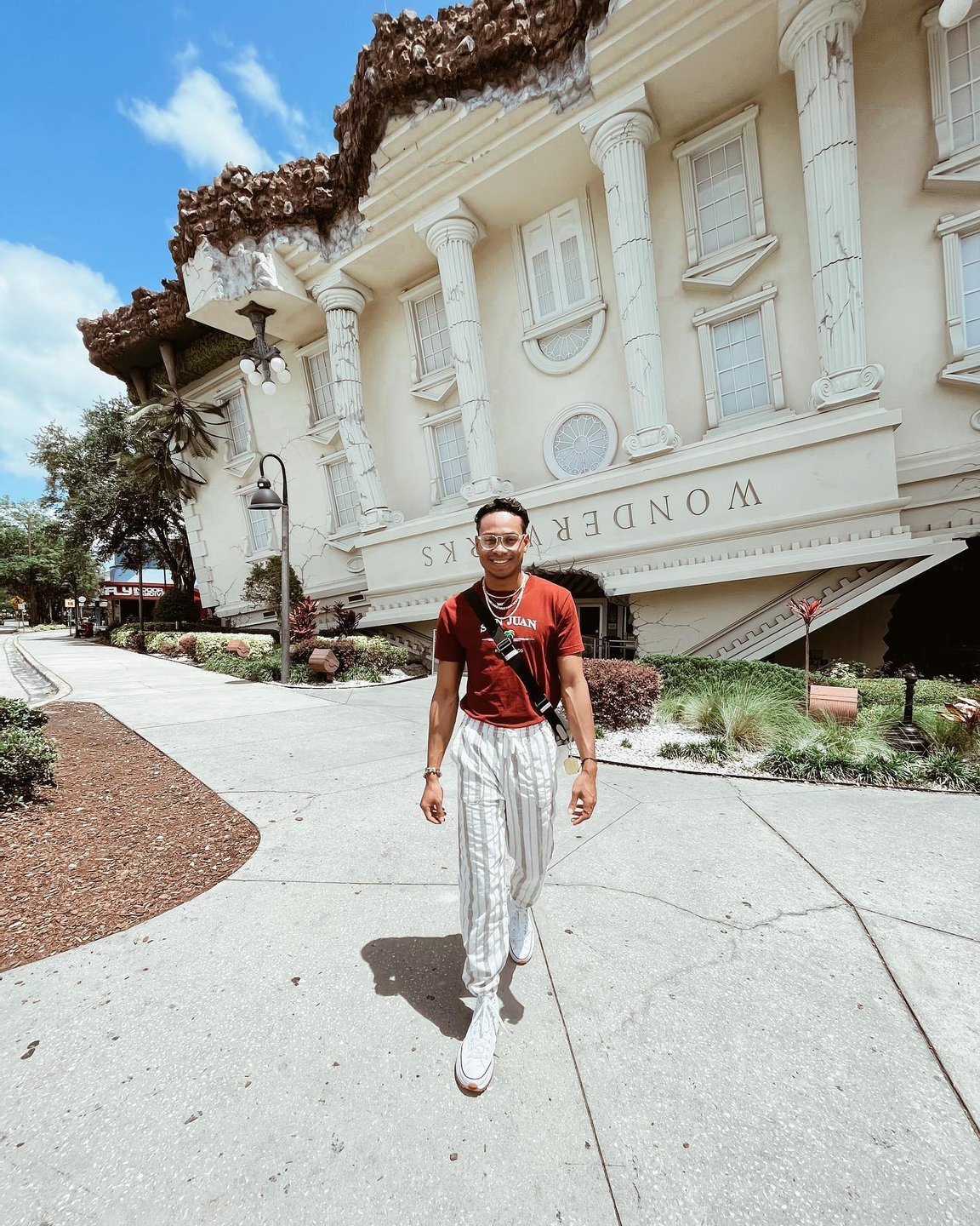 A person walks along a pathway in front of a whimsical, upside-down building labeled "Wonders." Lush greenery and palm trees surround the area.