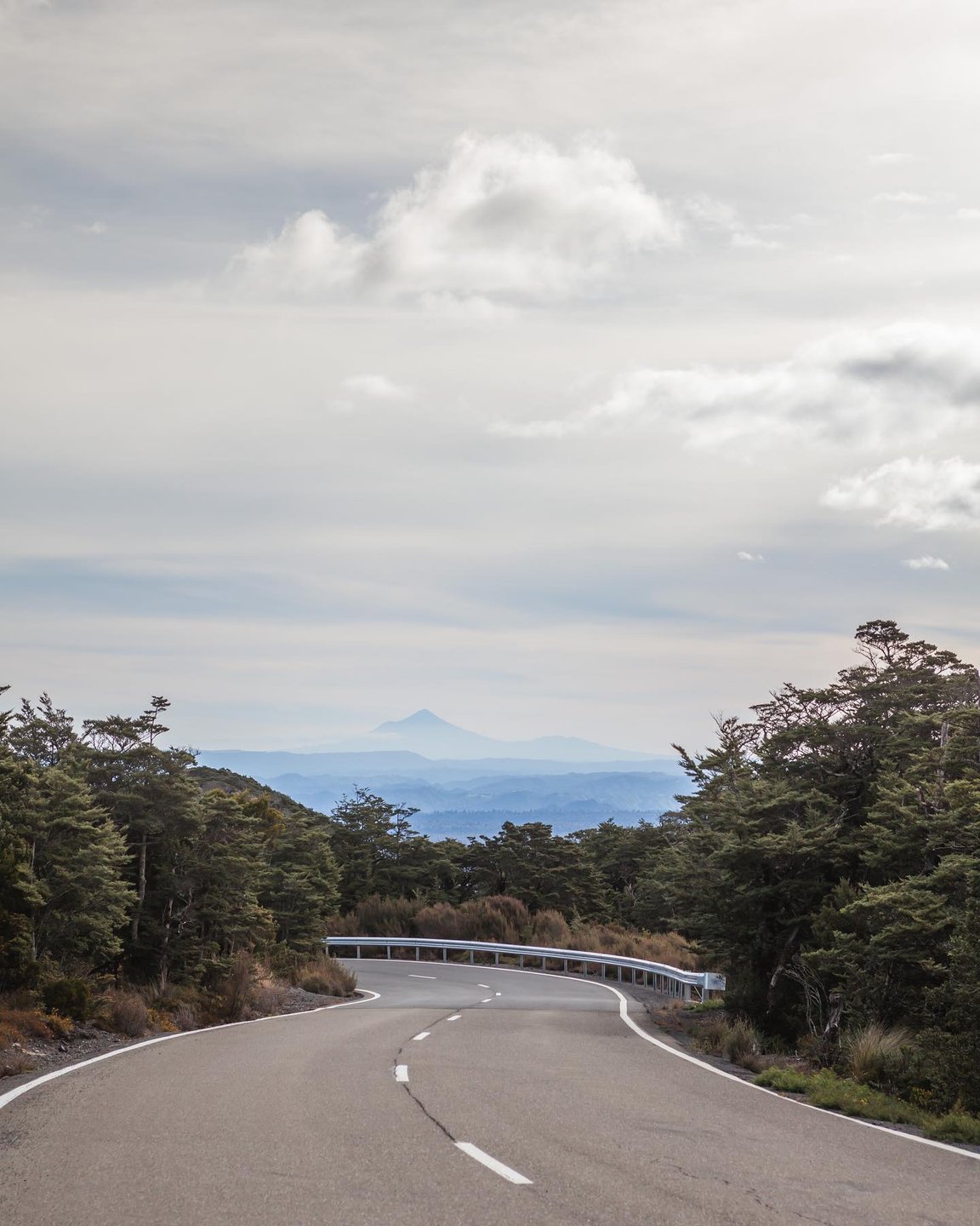 Winding road through lush greenery, leading to distant mountains under a cloudy sky. Ideal for scenic drives and exploration.