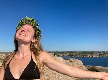 A woman in a black bikini stands on a rocky outcrop, wearing a leafy crown, with a scenic coastal view and clear blue sky behind her.
