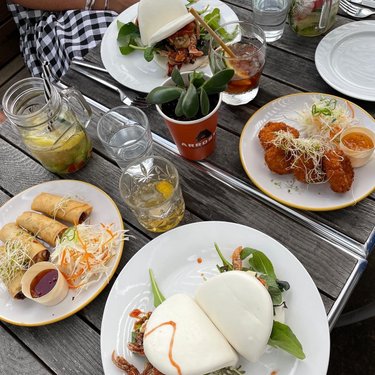 A table set with various dishes, including bao buns, spring rolls, and crispy chicken, surrounded by drinks and greenery.