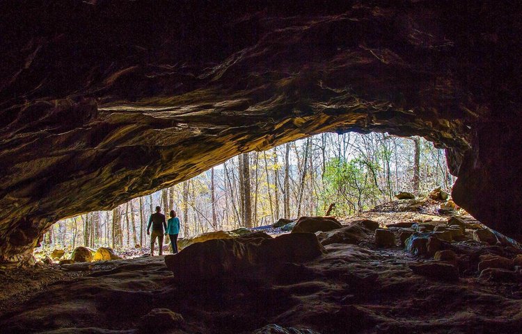A couple stands at the entrance of a cave, framed by rocky formations, overlooking a forest with trees and sunlight filtering through.