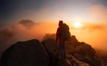 A hiker in a red jacket stands on rocky terrain, overlooking a misty landscape at sunrise, with soft clouds and distant mountains.