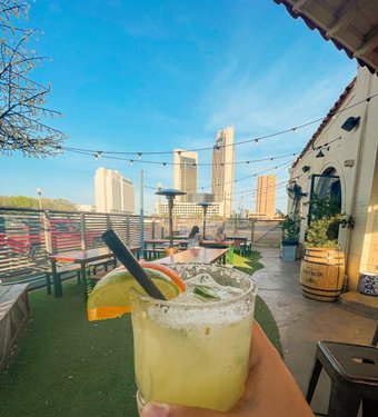 A hand holds a refreshing cocktail with a slice of orange, overlooking a rooftop patio with city skyscrapers and string lights.
