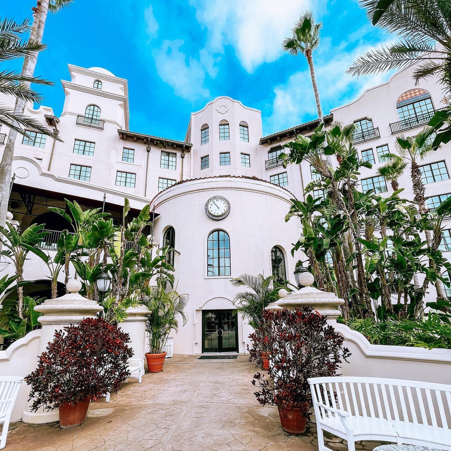 Elegant hotel entrance surrounded by lush greenery and palm trees, featuring a clock tower and inviting seating areas. Bright blue sky above.