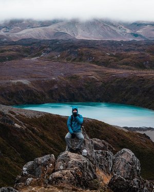 A person in a blue jacket sits on a rocky outcrop overlooking a turquoise lake surrounded by rugged mountains and misty clouds.