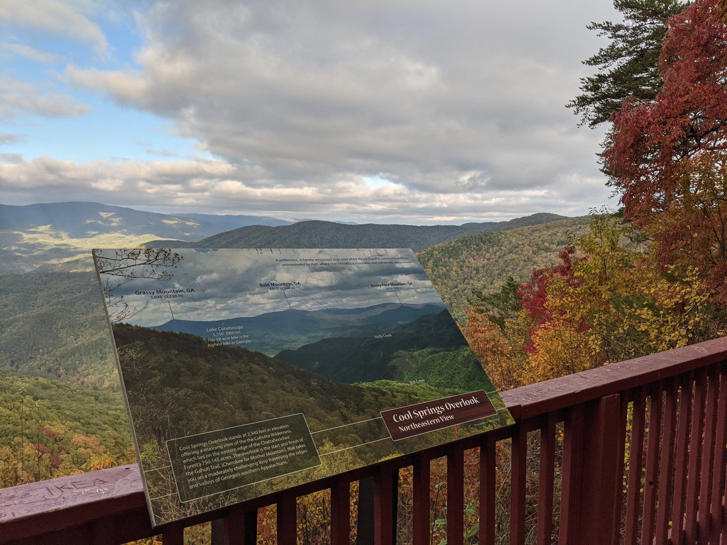 Scenic overlook with a wooden railing showcasing a panoramic view of rolling mountains and colorful autumn foliage under a cloudy sky.