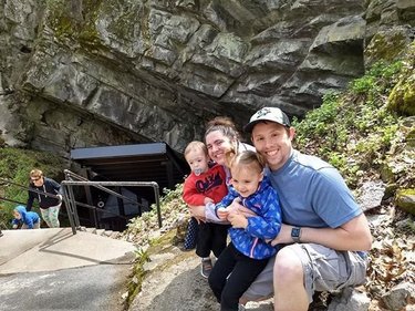 A family of four sits on a stone path near a rocky cliff, with two young children in their arms. A staircase leads to a cave entrance behind them.