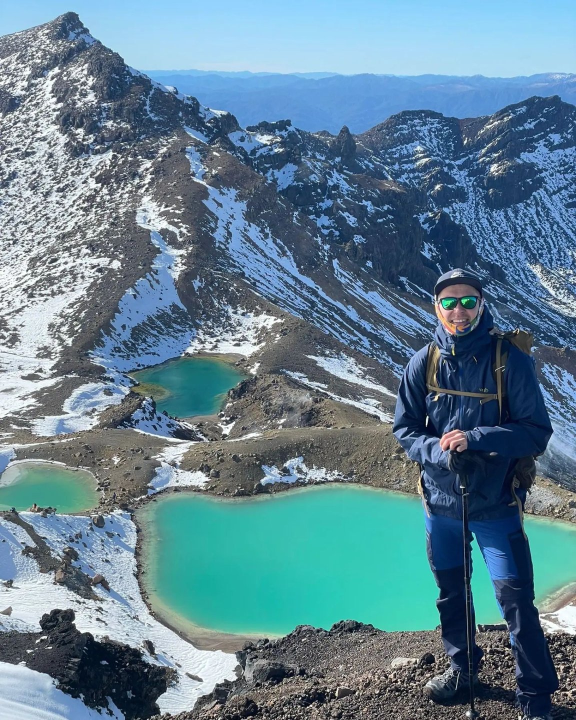 A hiker stands on a rocky mountain peak, overlooking turquoise glacial lakes surrounded by snow-capped peaks and clear blue skies.